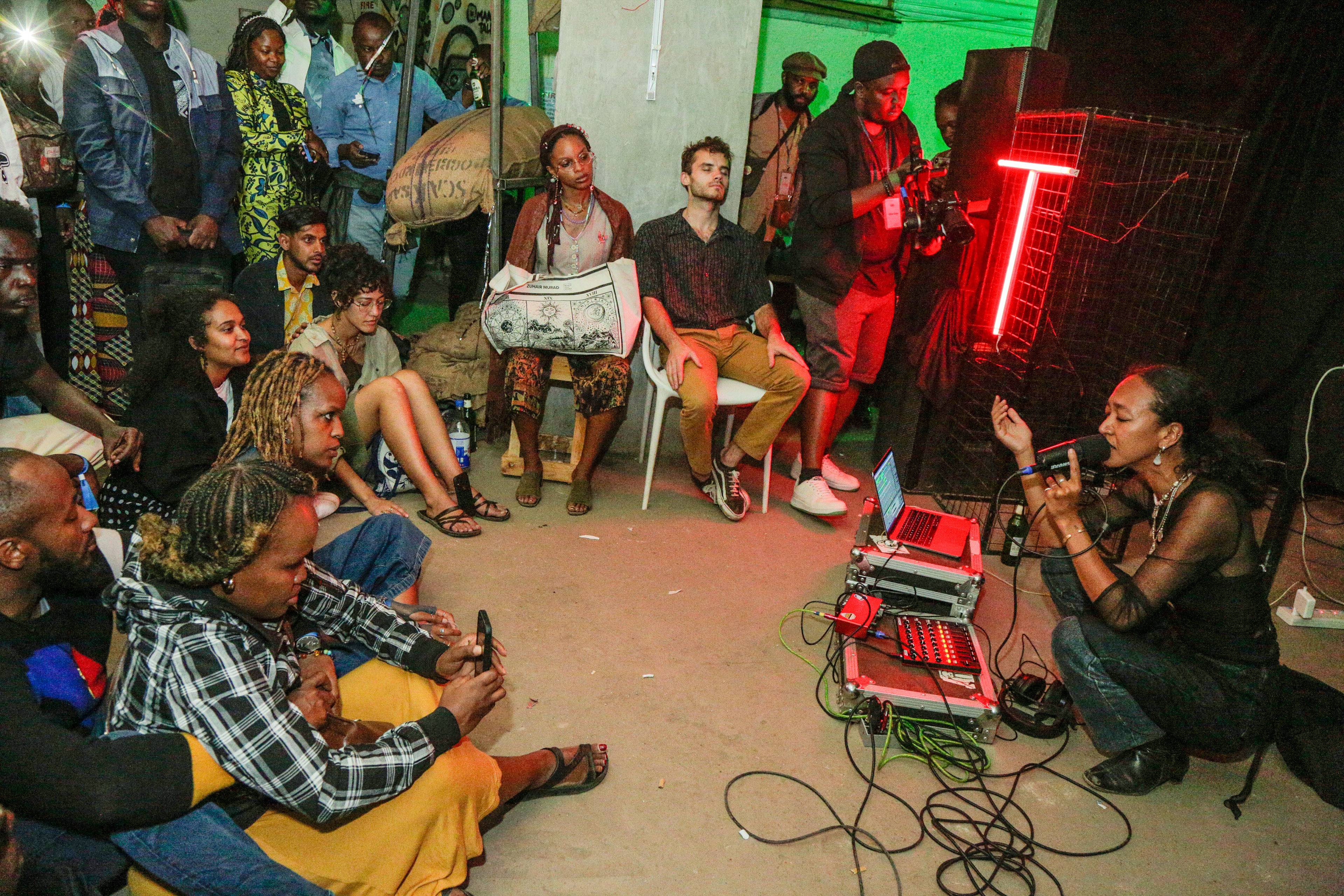 A crouching performer, singing with her eyes closed in front of an electronic set up for a small crowd.